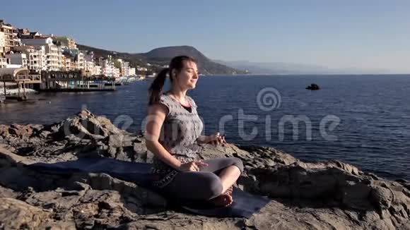 穿着运动服和耳机的女人坐在海上岩石上的莲花位置冥想听音乐瑜伽视频的预览图