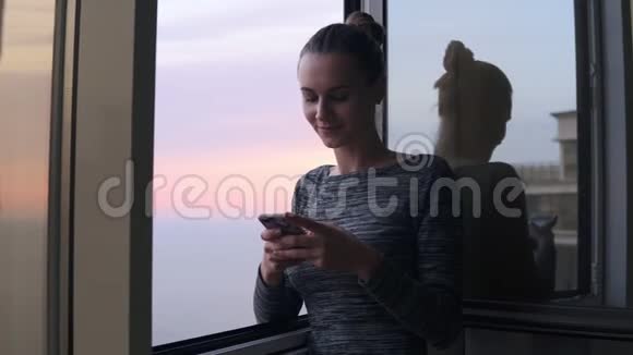 夕阳西下年轻迷人的女子站在敞开的窗户旁一边微笑着发短信视频的预览图