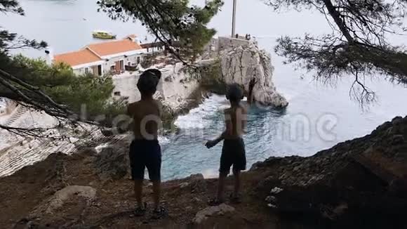 两个男孩站在高高的岩石上欣赏大海的后景两个哥哥站在高处的剪影视频的预览图
