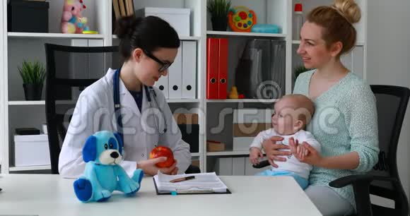年轻的女医生给母亲提供苹果和婴儿在办公室视频的预览图