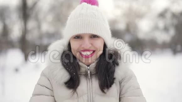 特写布鲁内特的头发年轻女性的肖像粉红色的嘴唇米色的羽绒服和帽子卷起雪微笑在雪中视频的预览图