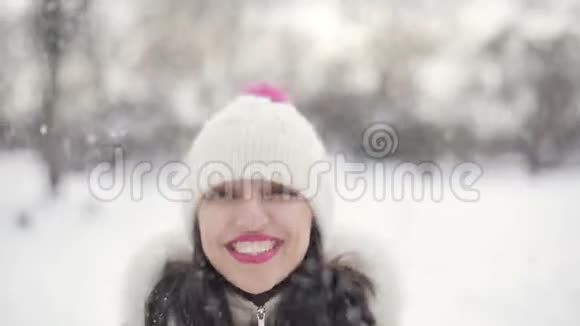 特写布鲁内特的头发年轻女性的肖像粉红色的嘴唇米色的羽绒服和帽子卷起雪微笑在雪中视频的预览图