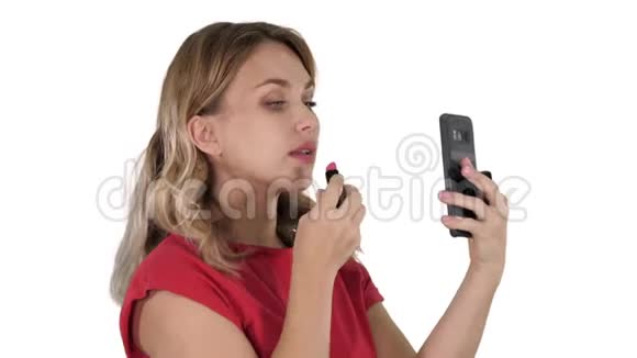 金发女人涂着口红在白色背景下看着手机视频的预览图