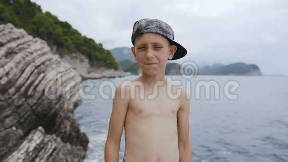 白人小男孩站在岩石上微笑着看着相机在海上背景下竖起大拇指夏季视频的预览图