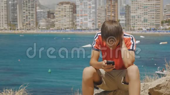 白人少年坐在石头上看着海上摩天大楼天际线背景上的智能手机屏幕视频的预览图