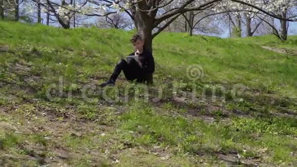 可爱的男孩在公园的草地上坐在花树下写诗视频的预览图