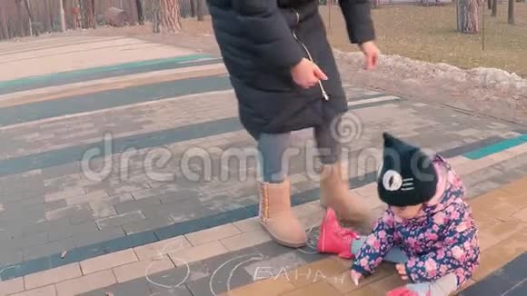 一个穿着五颜六色的粉红色夹克的小女孩和一位年轻的母亲在操场附近的人行道上画着粉笔画视频的预览图