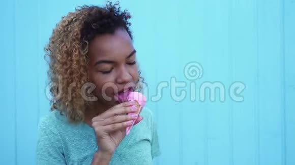 年轻的黑人女孩吃着粉红色的冰淇淋微笑着看着蓝墙背景视频的预览图