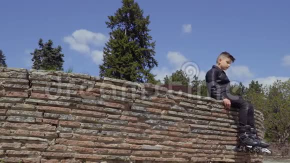 穿皮夹克和溜冰鞋的男孩坐在公园的混凝土墙上视频的预览图