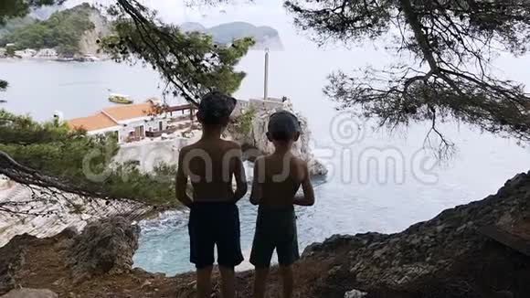两个男孩站在高高的岩石上欣赏大海的后景海滩度假两个哥哥站着的轮廓视频的预览图