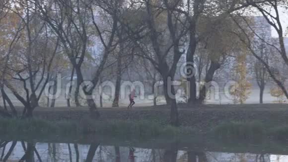 早晨一位年轻女子在浓雾中沿着河岸在一个城市公园慢跑任何天气的健康生活方式视频的预览图