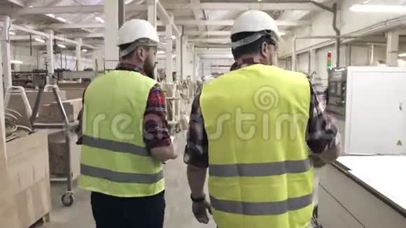 在一家家具厂两名工人穿着绿色背心和头盔在机器间交谈视频的预览图