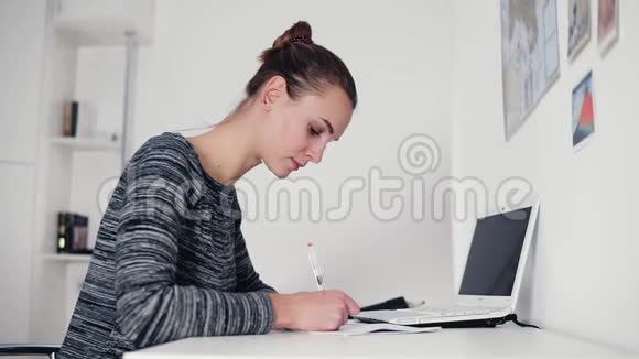 美丽的年轻女子坐在桌子旁用钢笔写一封信打开桌上的笔记本电脑女商人视频的预览图