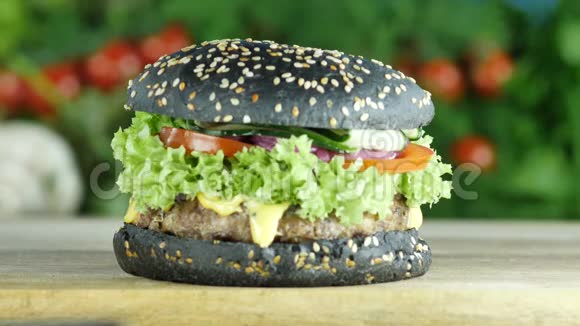 在绿色和蔬菜的模糊背景上带有黑色面包和芝麻的汉堡或汉堡包在木板上视频的预览图