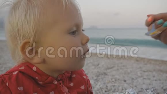 在海边背景上用勺子特写女性手喂养金发婴儿视频的预览图