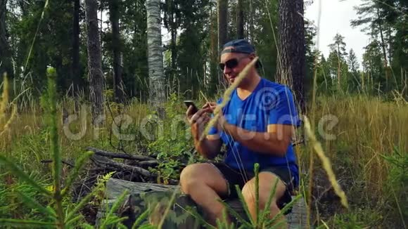 一位戴着太阳镜和棒球帽的游客坐在一棵倒下的树上用信使在电话里交流视频的预览图