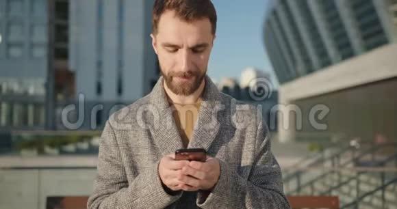 城市里穿着外套在手机上浏览和聊天的留胡子的时髦千禧男子的肖像手持射击视频的预览图