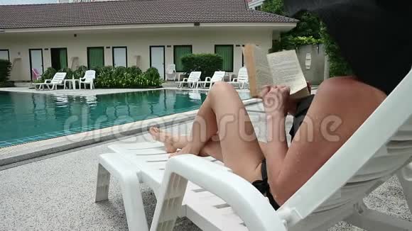 年轻漂亮的女人在游泳池附近读一本书苗条的女孩坐在躺椅上视频的预览图