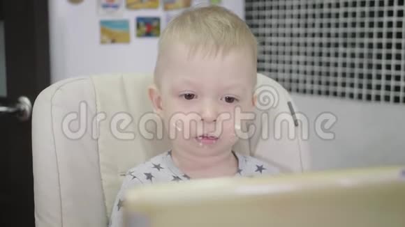 可爱的小男孩在家吃粥吃早餐同时在平板上看卡通儿童和技术视频的预览图