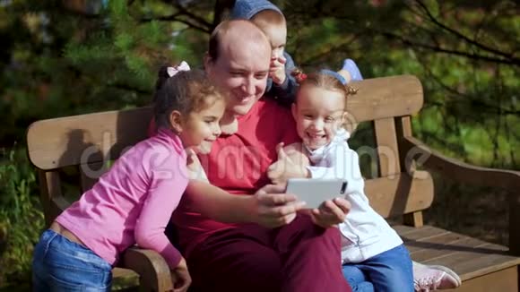 一个带孩子的男人坐在长凳上的智能手机上拍照幸福的一家人在公园里自拍家庭视频的预览图