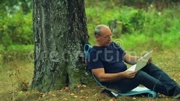 一个戴眼镜的人坐在公园的一棵树下用笔记本电脑工作视频的预览图