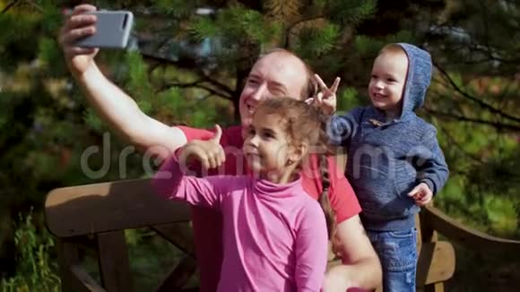 一个带孩子的男人坐在长凳上的智能手机上拍照幸福的一家人在公园里自拍家庭视频的预览图