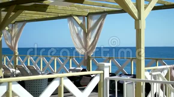 露台上有木制框架和白色窗帘以及海边沙滩上的躺椅在夏天阳光明媚的日子视频的预览图