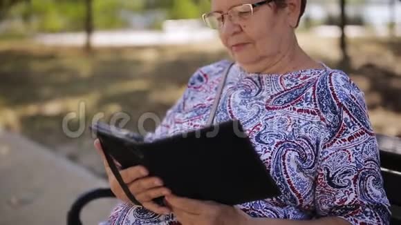 公园里戴眼镜的老妇人坐在长凳上看一本电子书视频的预览图