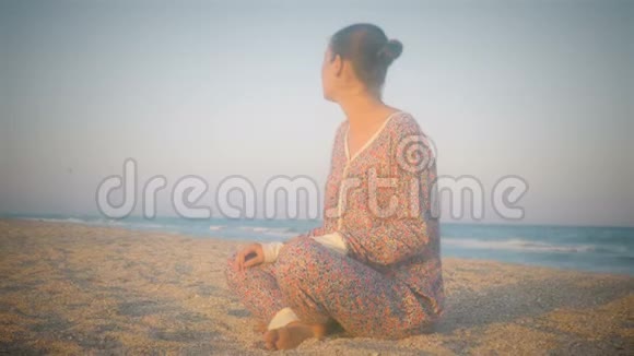女孩坐在海滩上摆出沉思的姿势微笑着享受海风和日落视频的预览图