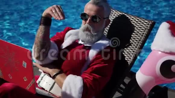 有趣的圣诞老人用笔记本电脑坐在躺椅上开玩笑视频的预览图