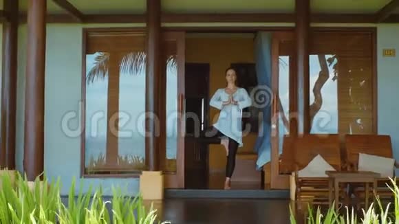 女人在树的位置练习瑜伽冥想和伸展在巴厘岛海滩上的平房门廊上大自然的声音视频的预览图
