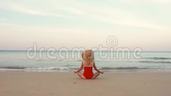 穿着红色泳衣和帽子的快乐女旅行者在完美的海滩上摆出莲花的姿势视频的预览图