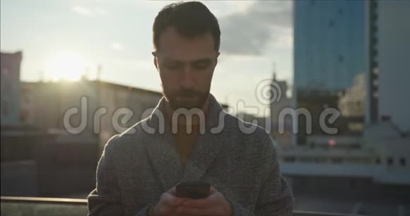 城市里穿着外套在手机上浏览和聊天的留胡子的时髦千禧男子的肖像手持射击视频的预览图