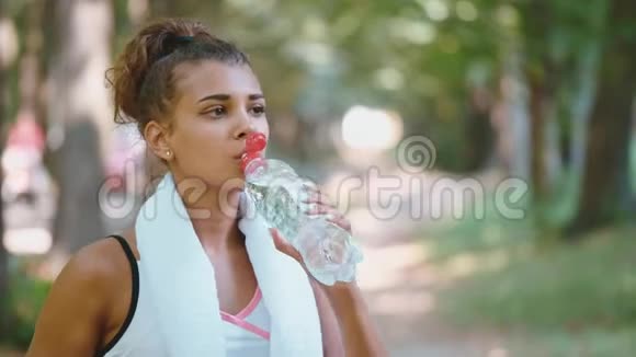 健康的女跑步者肩膀上有一条白色毛巾早上慢跑时用塑料瓶喝水视频的预览图