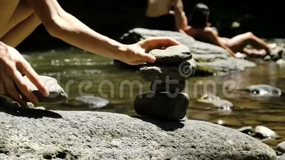 在游客的背景下女孩在一条山河上的鹅卵石上放了一个凯恩的特写镜头塔视频的预览图