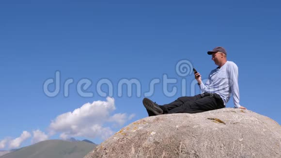 成年男子坐在大石头上在山区旅行中使用手机视频的预览图