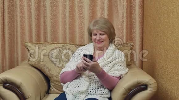 快乐的老妇人坐在一张舒适的椅子上拿着一个小玩意在家里休息迷人的微笑女士用触摸视频的预览图