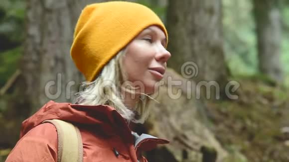 戴着一顶带背包的黄色帽子的金发美女在针叶林里四处张望女旅行者慢吞吞的肖像视频的预览图