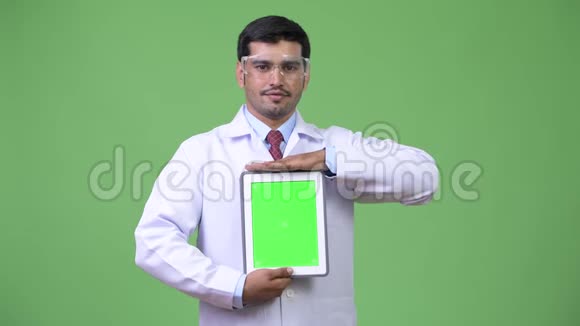 年轻帅气的波斯男医生戴着护目镜展示数码平板电脑视频的预览图