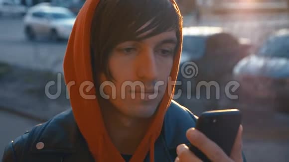 英俊的年轻人在阳光城市街道城市背景穿红色连帽衫的人用智能手机日落背景视频的预览图