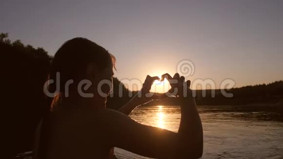 快乐的女孩晚上在夕阳对面的河边洗澡展示着心的手视频的预览图