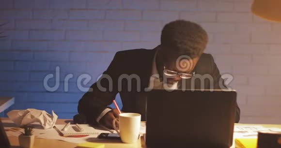 年轻的黑人商人带着笔记本电脑和文件在夜间办公室工作他在文件上签字商务视频的预览图