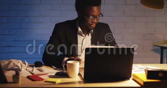 年轻的黑人商人带着笔记本电脑和文件在夜间办公室工作他在文件上签字商务视频的预览图
