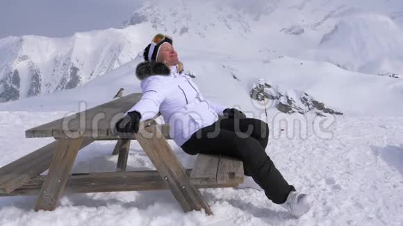 在阳光明媚的日子里坐在长凳上女人滑雪者很高兴在山上放松视频的预览图