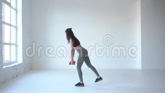 心血管教程这个年轻的运动女孩正在用哑铃做蹲在白色的工作室里抬起腿4k视频的预览图