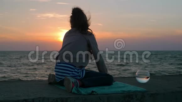 小女孩坐在沙滩上看美丽的日落鱼缸里养着宠物鱼在室外慢动作视频的预览图