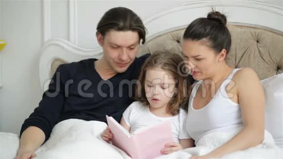 漂亮的小女孩正试图和她的父母躺在床上看书美丽的孩子正在用手指朗读视频的预览图