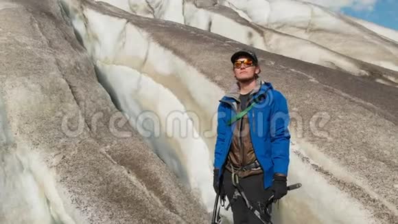 一位年轻的登山者戴着墨镜戴着帽子和棉条站在一座巨大的冰川上山脉上有巨大的裂缝视频的预览图