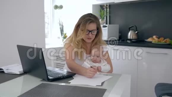 多任务妈妈抱着孩子在餐桌上工作在厨房跳儿子的背景下视频的预览图