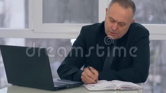 办公室年长的商人用笔记本电脑工作在笔记本上记笔记视频的预览图